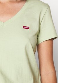 Jasnozielona bawełniana koszulka z dekoltem w serek i krótkim rękawem. Posiada małe czerwone logo Levis na lewej stronie klatki piersiowej. Gładka faktura.