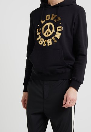 Persona con una sudadera negra con capucha, con un símbolo de paz dorado y texto "LOVE MOSCHINO", combinada con pantalones negros con rayas laterales blancas.