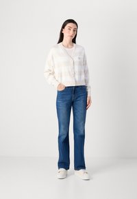 En kvinne står selvsikkert i en stripete cardigan og blå jeans, med en nøytral bakgrunn som fremhever hennes uformelle antrekk.