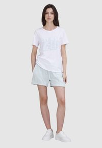 T-shirt en coton blanc avec le texte bleu clair "TRUE JEAN BABY" ; associé à un short vert clair et des baskets blanches.