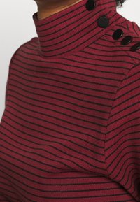 Suéter de cuello alto a rayas rojas y negras con un alto cuello y botones negros en el hombro, exhibido en una tela suave y texturizada.