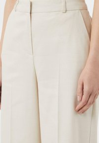 Jasno beżowe spodnie wykonane z teksturowanej tkaniny, charakteryzujące się dopasowanym krojem, zaszewkami i paskiem z pętlami na pasek. Ręka opiera się na boku.