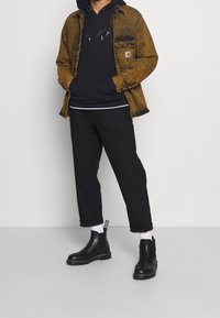 Brun denimjacka, svart huvtröja, svarta byxor och svarta chelsea-boots. Vita strumpor synliga med logotyp.