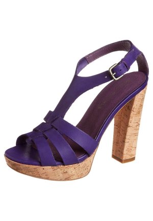 Lilla sandal med høje hæle, korksål og chunky hæl. Har brede stropper og en ankelspænde for sikker pasform. Glat tekstur.