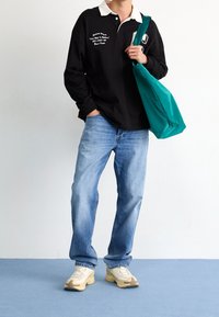 Polo negro de manga larga con cuello blanco y bordado, acompañado de jeans de color azul claro y zapatillas color crema. Bolsa de mano verde colgada del hombro.