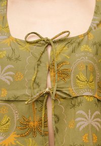 Κοντινό πλάνο πράσινου υφάσματος με κίτρινο και πορτοκαλί τροπικό σχέδιο φοίνικα και λουλουδιών, δεμένο σε δύο κάθεντες φιόγκους πάνω σε γυμνό δέρμα.