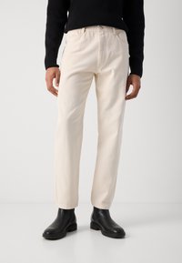 Pantalones de color crema con un corte recto, hechos de una tela suave. Presentan un cierre con botón y bolsillos laterales. Combinados con botas negras hasta el tobillo.