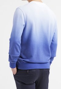 Hombre vistiendo un sudadera azul ombré de manga larga y pantalones negros, de espaldas con las manos en los bolsillos contra un fondo blanco.