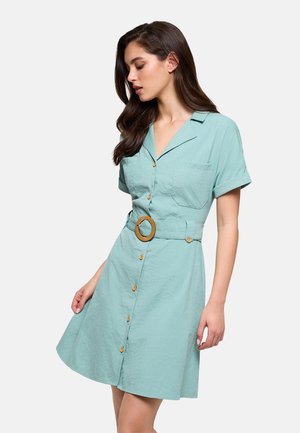 Mujer con un vestido azul claro de manga corta con botones, con cuello y un cinturón ancho con una hebilla redonda.