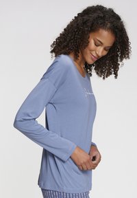 Long-sleeve light blue top made of soft fabric, featuring a rounded neckline and an embroidered detail on the front. Hem sits below the hips.