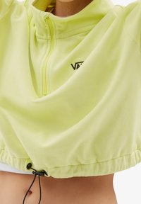 Pulóver amarillo claro con cremallera y diseño cropped. Cuenta con un dobladillo elástico en la cintura y un pequeño logotipo negro en el pecho. Textura suave.