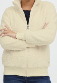 Persona vistiendo una chaqueta de cremoso peluche con cremallera sobre una parte superior oscura y jeans azules, con los brazos cruzados sobre el pecho.
