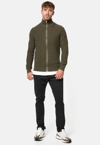 Suéter de punto acanalado verde oliva con cremallera y cuello alto, llevado sobre una camisa blanca, combinado con pantalones negros y zapatillas blancas.