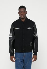 Fekete sportdzseki bőr ujjakkal, mellkason és a sleveken logók találhatók, valamint hímzett szöveg az elején. Farmernadrág is tartozik hozzá.