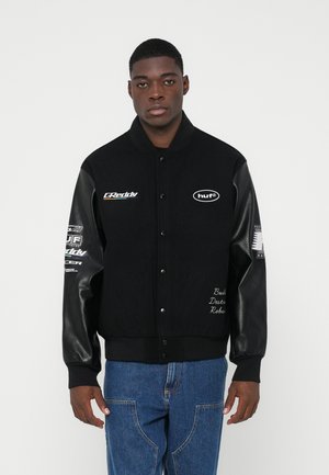 Melnā universitātes jakā ar ādas piedurknēm, kurai ir logotipi uz krūšu un piedurknēm, kā arī izšūta teksts uz priekša. Iekļautas džinsu bikses.