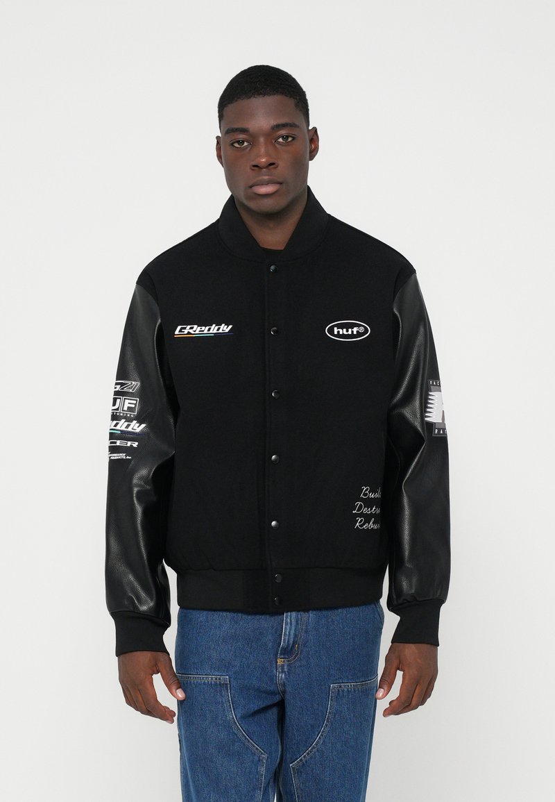 Fekete sportdzseki bőr ujjakkal, mellkason és a sleveken logók találhatók, valamint hímzett szöveg az elején. Farmernadrág is tartozik hozzá.