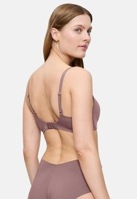 Γυναίκα με μακριά μαλλιά, φορώντας μωβ σουτιέν και αντίστοιχο ψηλόμεσο εσώρουχο, γυρισμένη προς τα πίσω και κοιτάζοντας πάνω από τον ώμο της.