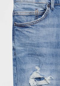 Ljust blå denimjeans med en liten framficka, orange sömmar, metalnitar och ett trasigt hål nära låret.