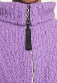 Pull en tricot lavande épais avec une texture côtelée, un col montant et une fermeture éclair argentée dotée d'un tirette noir.
