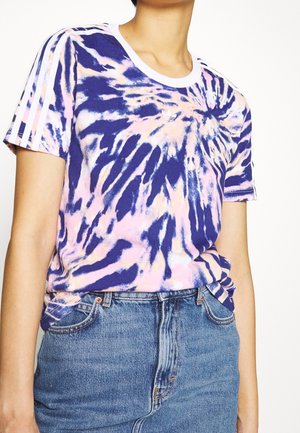 Osoba nosí modrobílorůžové tričko Adidas s tie-dye vzorem, bílé pruhy na rukávech a modré džíny s vysokým pasem.