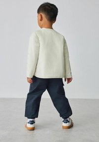 Weiße gesteppte Jacke, dunkelblaue Cargohose und weiße Turnschuhe mit dunkelblauen Akzenten. Kind von hinten gesehen, stehend auf einer hellgrauen Fläche.