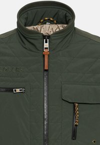 Grøn quiltet jakke med høj krave, frontlynlås, læder trækflig og flere lommer. Har kontrasterende syninger og hardware-detaljer.