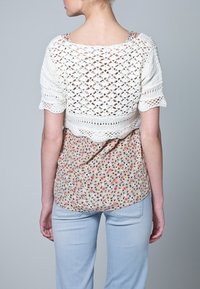 Bílý háčkovaný top s krátkým rukávem přes květovanou blůzu, doplněný světlými modrými džínami. Háčkování má krajkovou texturu a otevřený pletený vzor.