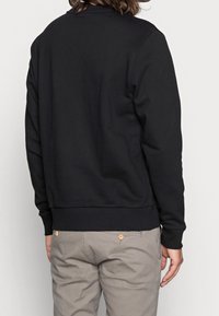 Henkilö, jolla on olkapituiset hiukset, pukeutuu mustaan pitkähiharaiseen sweatshirtiin ja vaaleanharmaisiin housuihin, näkyy selkäpuolelta yksinkertaista valkoista taustaa vasten.