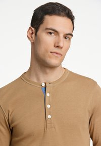 Brun långärmad henley-tröja med rundad halsringning, fem vita knappar framtill och en blå accent på vänster sida. Mjuk textur.