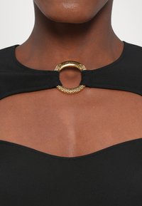 Zwarte kleding met ronde halslijn, met een gouden ronde hardware detail in het midden en textuuraccenten op de ring.