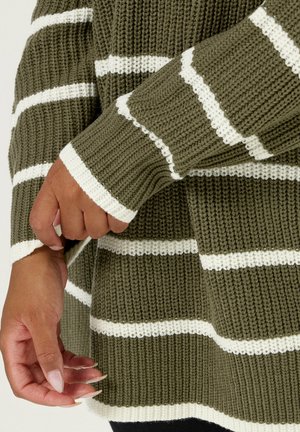 Pull en tricot vert olive avec des rayures horizontales blanches. Présente une texture côtelée et des manches larges. Le poignet montre une main agrippant le tissu.