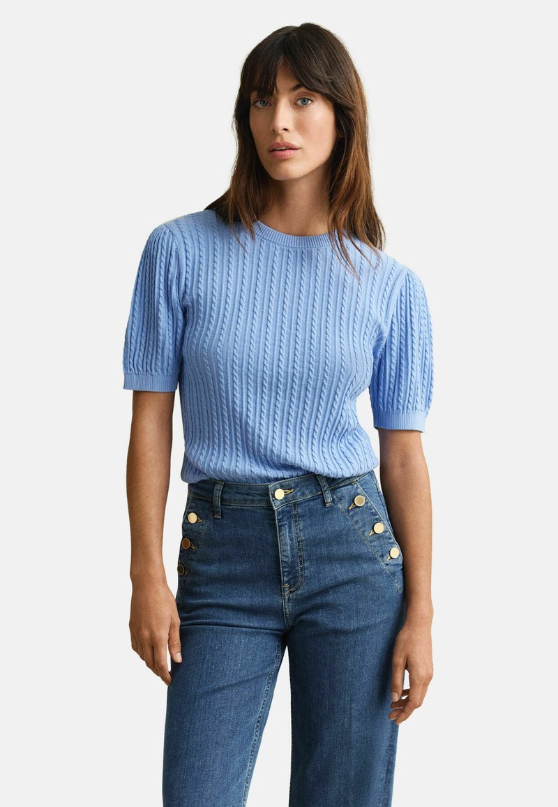 Femme portant un pull en maille torsadée bleu clair à manches courtes et un jean taille haute bleu foncé avec des boutons dorés sur les poches.