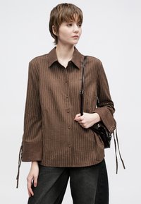 Ruda dryžuota ilgomis rankovėmis palaidinė su apykakle, su užsegimu mygtukais ir raišteliais ant riešų. Modelis laiko mažą tamsią rankinę.