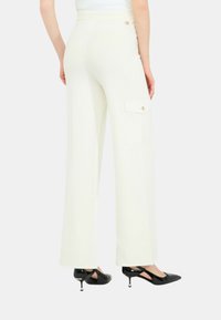 Pantaloni a gamba larga in crema con una texture liscia. Presentano una tasca laterale tipo cargo e un sottile accento dorato in vita. Abbinati a tacchi neri.