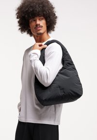 Borsa a spalla nera imbottita con una texture morbida e una forma arrotondata, caratterizzata da un dettaglio del logo. Indossata da una persona con una camicia bianca a maniche lunghe.