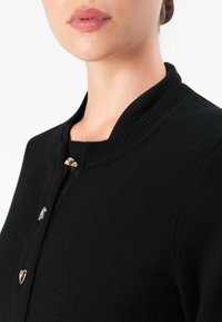 Kvinna iklädd en svart stickad kofta med tre distinkta gyllene knappar formade som ett skal, en cirkel och ett hjärta nära kragen.