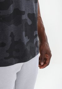 Camiseta sin mangas de camuflaje negra, con una textura suave y un ajuste holgado. Pantalones deportivos grises con puños acanalados visibles en la parte inferior.