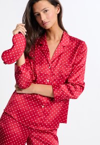 Conjunto de pijama de satén rojo con patrones de corazones blancos. Presenta una camisa abotonada de manga larga, bolsillo y pantalones a juego. Incluye una máscara para los ojos en forma de corazón.