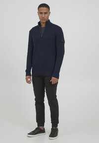 Pullover lavorato a maglia blu navy con colletto a mezzo zip, caratterizzato da maniche lunghe e un motivo testurizzato. Abbinato a pantaloni neri e sneakers nere.
