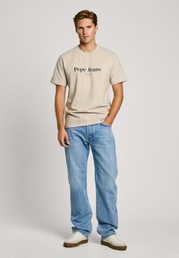 Gaiši beidzīgi kokvilnas T-krekls ar "Pepe Jeans" logotipu, apvienots ar gaiši ziliem džinsiem un baltām kedām. Brīva piegriezuma, īsās piedurknes.