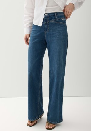 Persoon die blauwe wijde jeans, wit overhemd en open sandalen draagt, staand met één hand in de heup tegen een effen achtergrond.