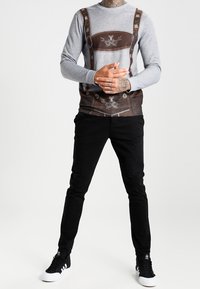 Long-sleeve gray shirt with a brown faux lederhosen print, black pants, and black sneakers, featuring white soles and laces.