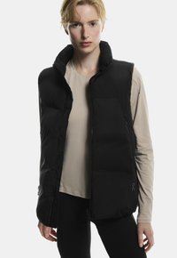 Sort pufvest med høj krave, lynlås og forlommer. Blød tekstur med en glat overflade, parret med en lys beige top.