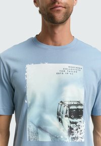 Muž v svetlom modrom tričku s grafikou terénneho vozidla jazdiaceho po zasneženej alebo zamrznutej ploche a textom "Northern Collective Tom Tailor ESTD 1962".