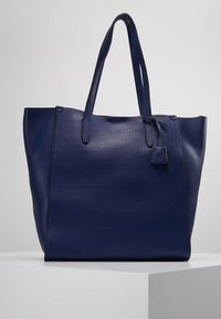 Laivastonsininen nahkainen olkalaukku, jossa on teksturoitu pinta, kaksinkertaiset kahvat ja kiinnitetty pieni merkki. Leveä avoin yläosa helppoon pääsyyn.