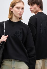 Δύο νέοι ενήλικες φορούν μαύρα φούτερ με το κείμενο «EST 1978», ο ένας κρατά μια μαύρη τσάντα, και οι δύο κοιτούν προς ελαφρώς διαφορετικές κατευθύνσεις.