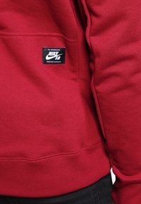 Röd sweatshirt i mjukt tyg, med en stor känguruficka, ribbade muddar och en svart Nike SB-logetikett på sidan.