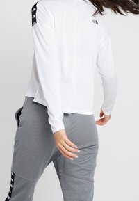 Person i vit långärmad skjorta och gråa sweatpants med svarta logostripes, i rörelse mot en enkel bakgrund.