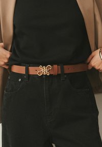 Cinturón de cuero color marrón claro con una hebilla dorada decorativa de diseño ornamentado. Se lleva sobre pantalones vaqueros negros y cuenta con múltiples orificios para ajustar.