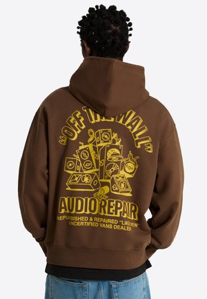Hombre con cabello rizado lleva una sudadera marrón con capucha con un gráfico amarillo de altavoces apilados y el texto "Off The Wall Audio Repair" en la espalda.